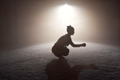En dansare sitter på huk med armarna sträckta framför sig. Rummet har dämpad belysning med en strålkastare som är riktad mot dansaren. Vi ser hennes siluett.